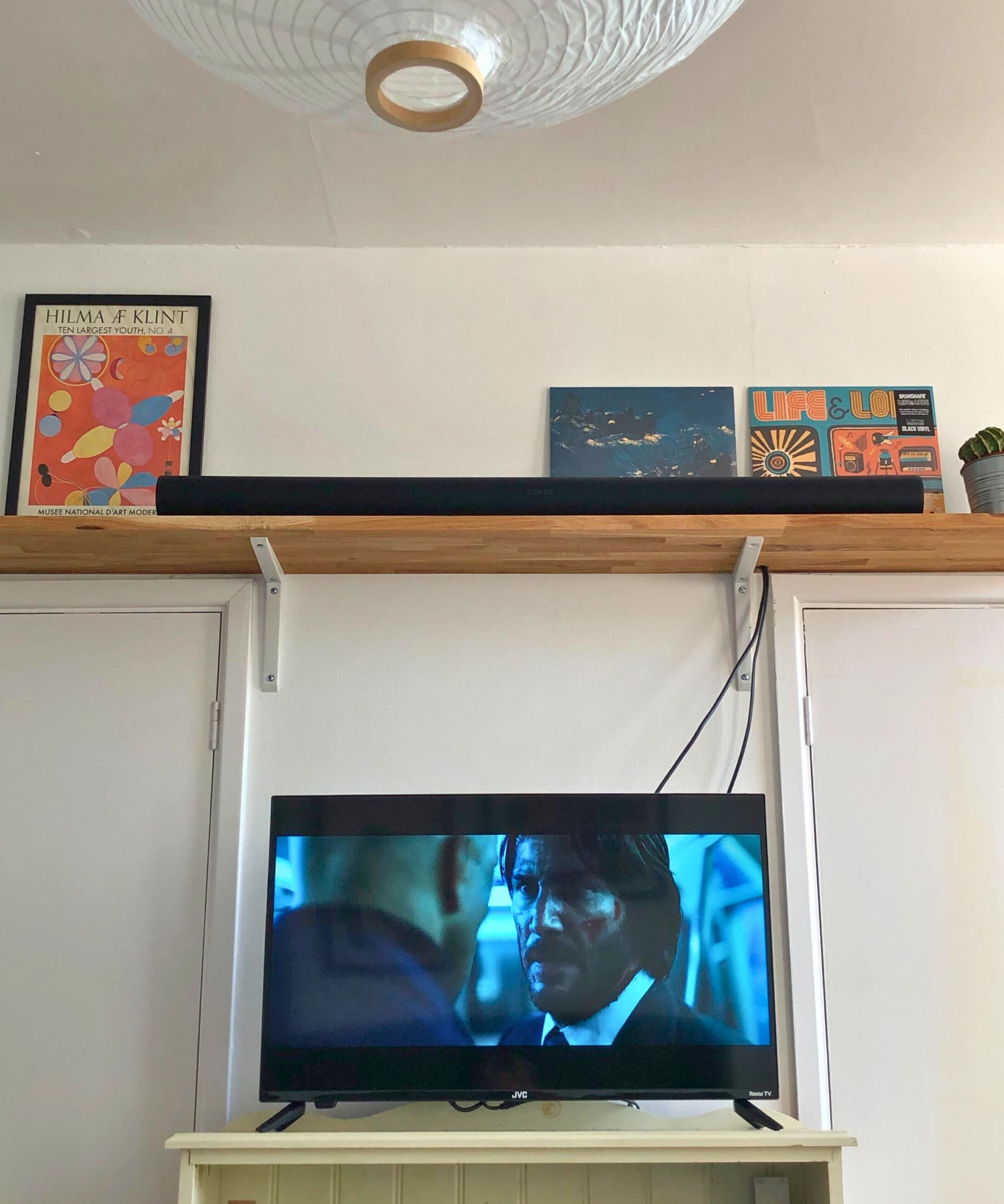 Sonos Arc Ultra soundbar in Dan Fauzi's home, on wooden shelf with cactus plant, above TV showing John Wick: Chapter 2. A white lampshade is pictured in the top left corner, above two prints on the wall.