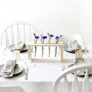 A wodden rack holds individual purple flowers in the centre of a white dining table setting in a white room.