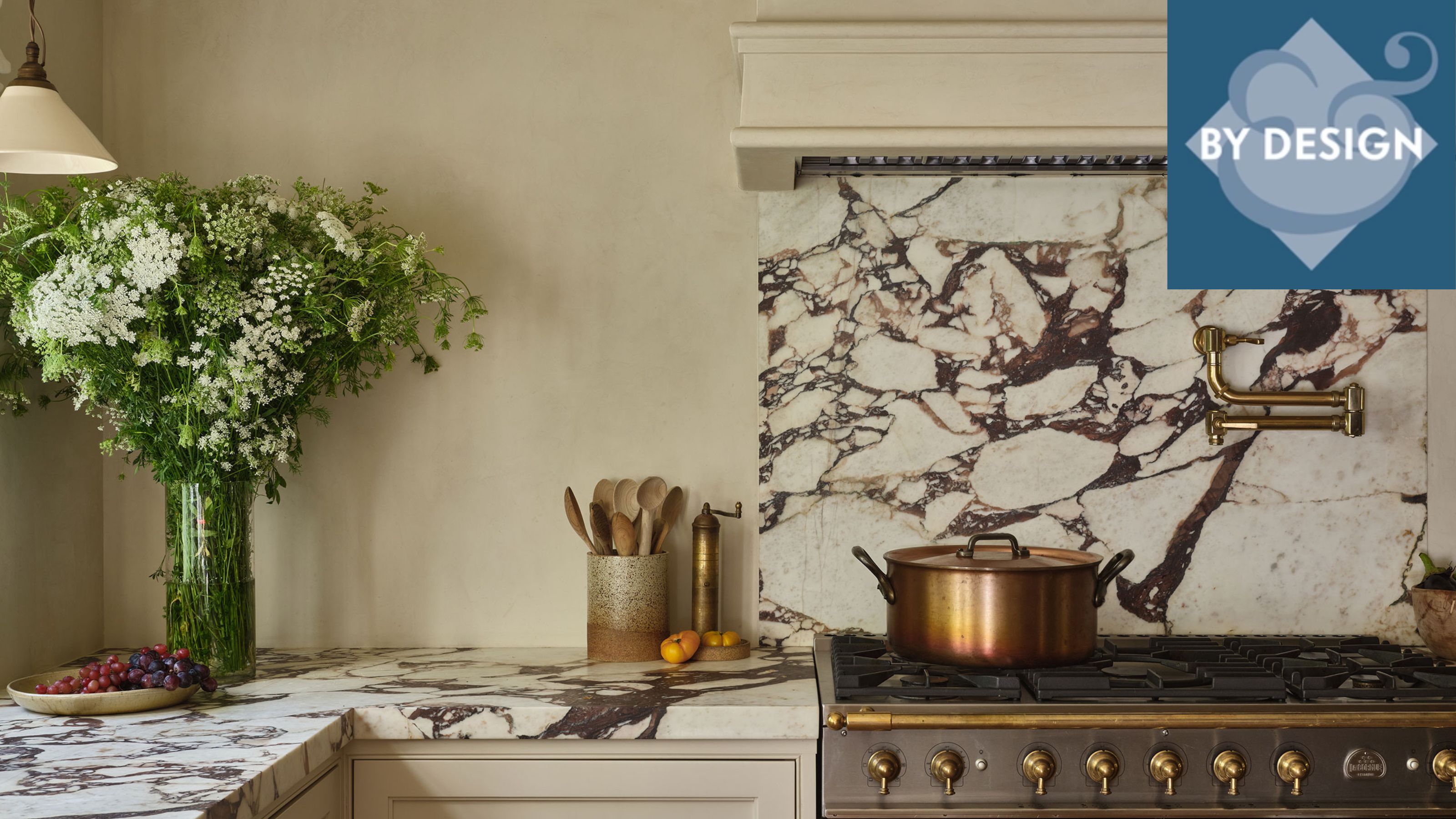 Close up of kitchen counter with marble countertop and backsplash. Stove with copper pots on top, counter decorated with flowers and untensils