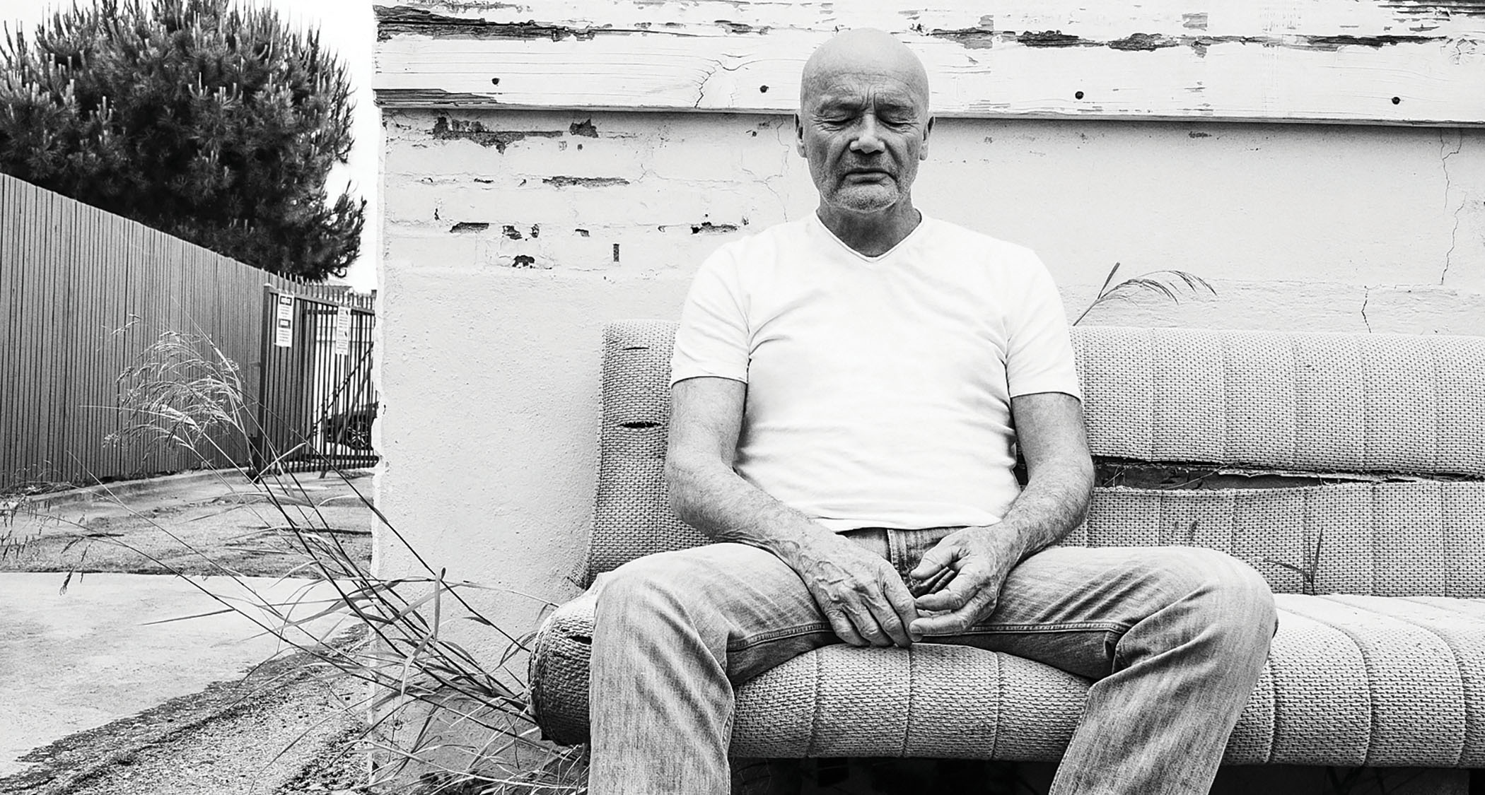 A black-and-white portrait of Creed Bratton sitting on a sofa outdoors.