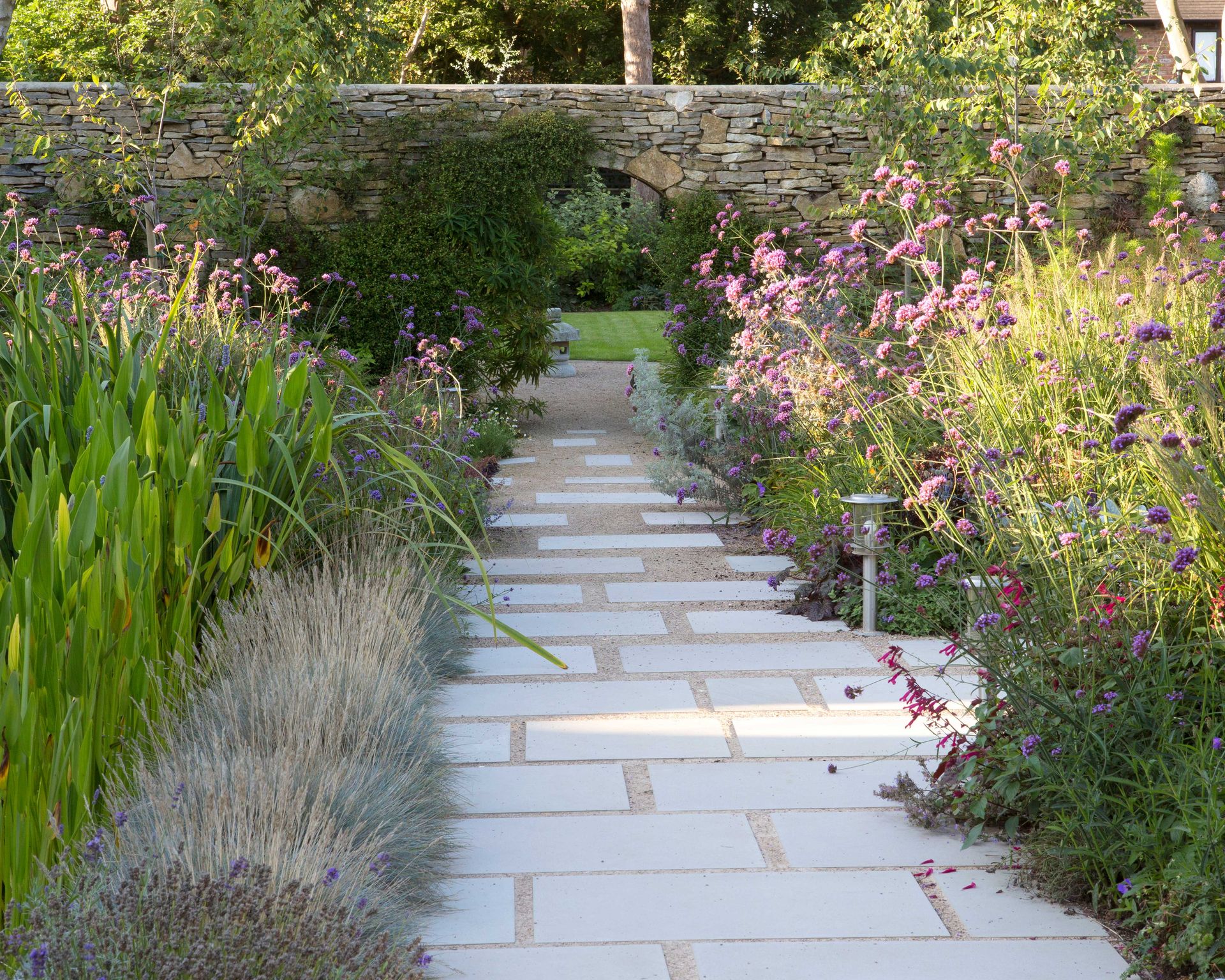 garden path with mixed smooth slabs and gravel through densely planted borders towards doorway in wall