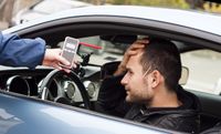 A white man with a beard sits in the driver's seat of a car clutching his forehead. A hand holds a breathalyzer test toward him