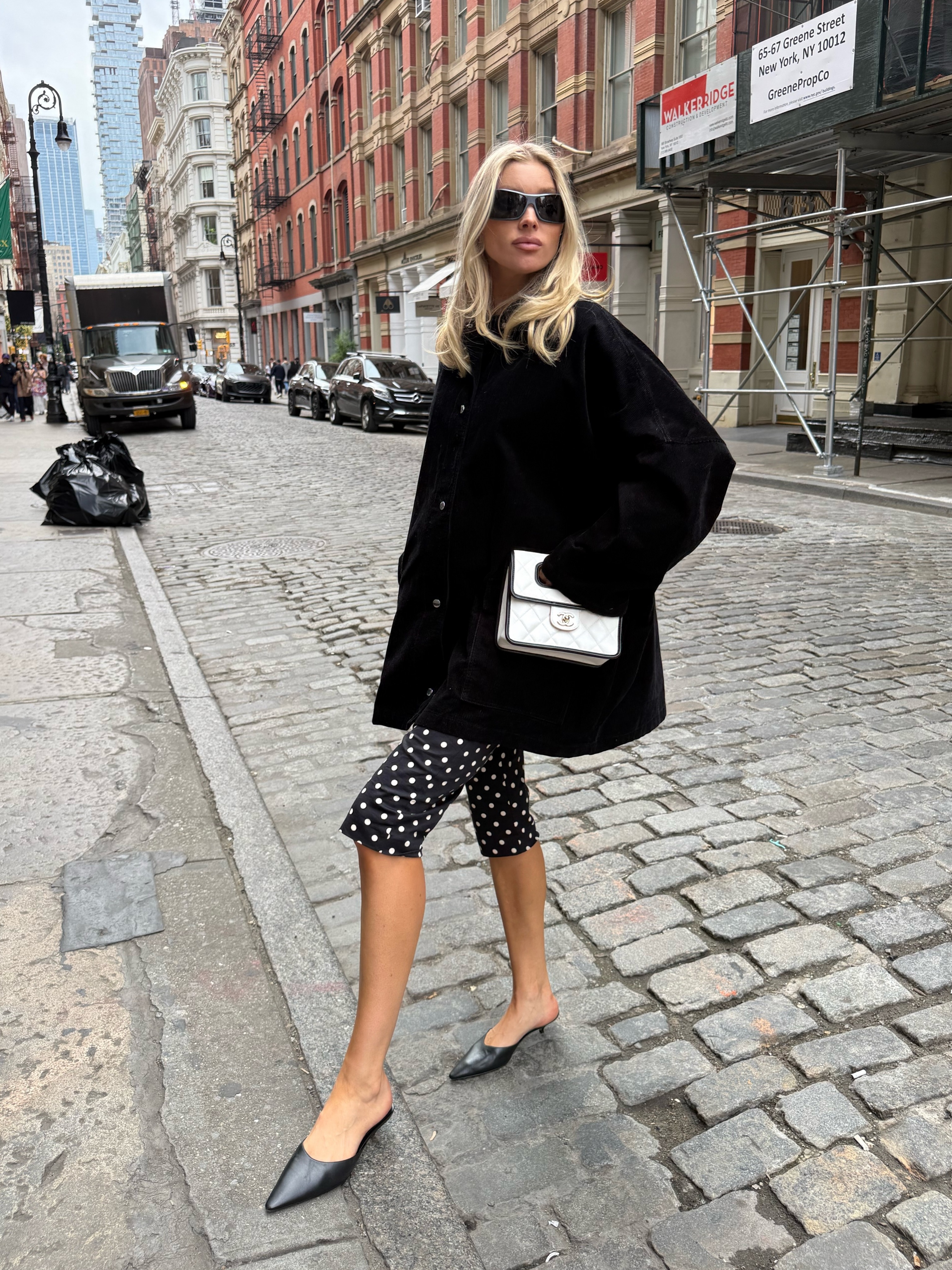 Elsa Hosk poses for a photo wearing polkadot capri pants with pointed-toe heeled mules and a black jacket.