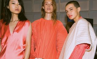 Models wear pleated blouses and coats in coral, cream and a coral silk dress