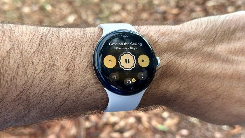 A photo of the Google Pixel Watch 4 on a wrist outdoors, showing music playback controls for a Pandora playlist.