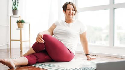 Woman performing seated twist (Ardha Matsyendrasana) yoga pose in domestic setting looking at an open laptop on the floor