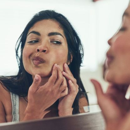 Woman looking in the mirror and popping a pimple on her cheek - how to pop a pimple