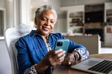 Older woman smiling and looking at cell phone while she sits at dining room table.