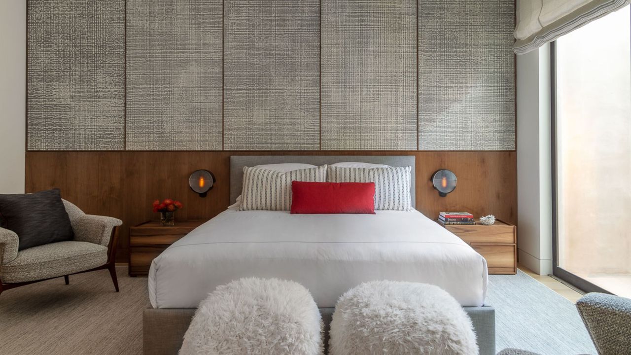 japandi style bedroom with textured wallpaper and timber panelling behind bed, an armchair, timber side tables, a gray upholstered bedframe with white sheets, striped and red pillows, and two fluffy ottomans at the end of the bed