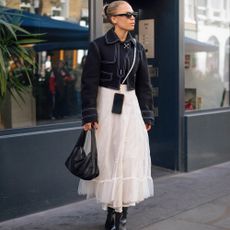 Woman wears cropped denim jacket in a deep wash and voluminous white tulle maxi skirt. She also wears black sunglasses, handbag, and boots.