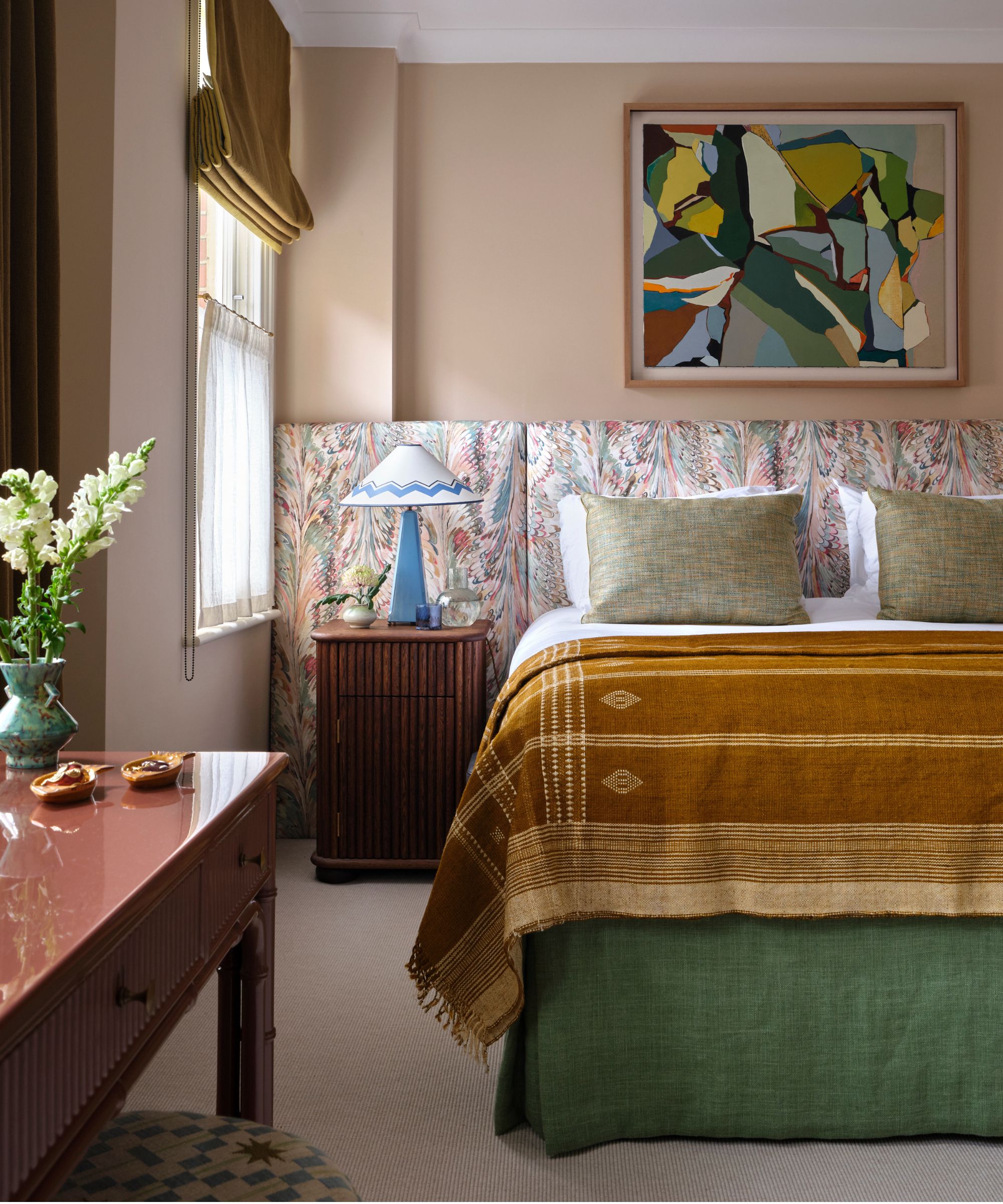 Bedroom with plaster pink walls, fabric headboard, bed with mustard quilt, mahogany desk and ottoman, and abstract colourful painting above bed