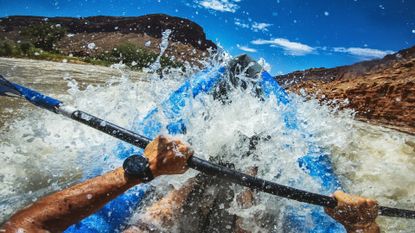 A man's hands hold an oar while he navigates rough water in a kayak.