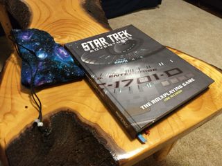 A game book sits on a wooden, tree-trunk-like table next to a galaxy-patterned dice bag.