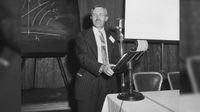 a man stands during a presentation wearing a suit and tie.