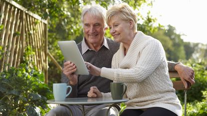 A smiling older couple sit outside and look at a tablet together.