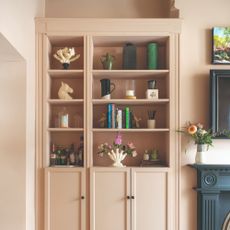A built-in bookcase in a living room colour drenched in soft pink with the shelves filled with decorative objects