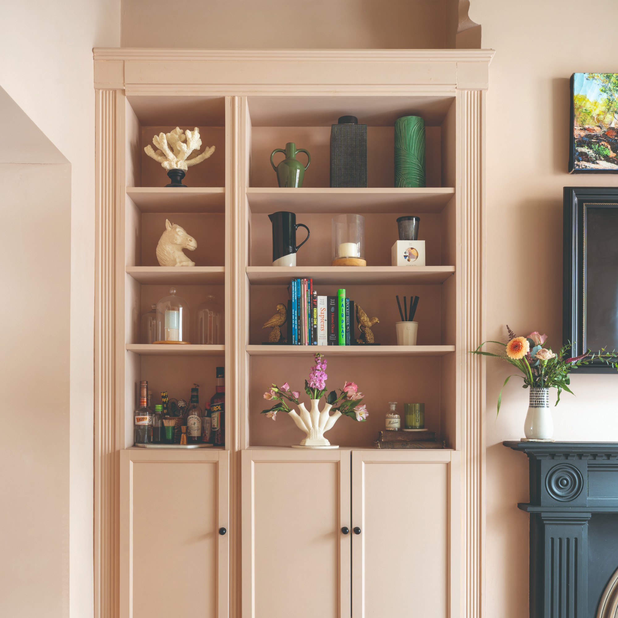 A built-in bookcase in a living room colour drenched in soft pink with the shelves filled with decorative objects