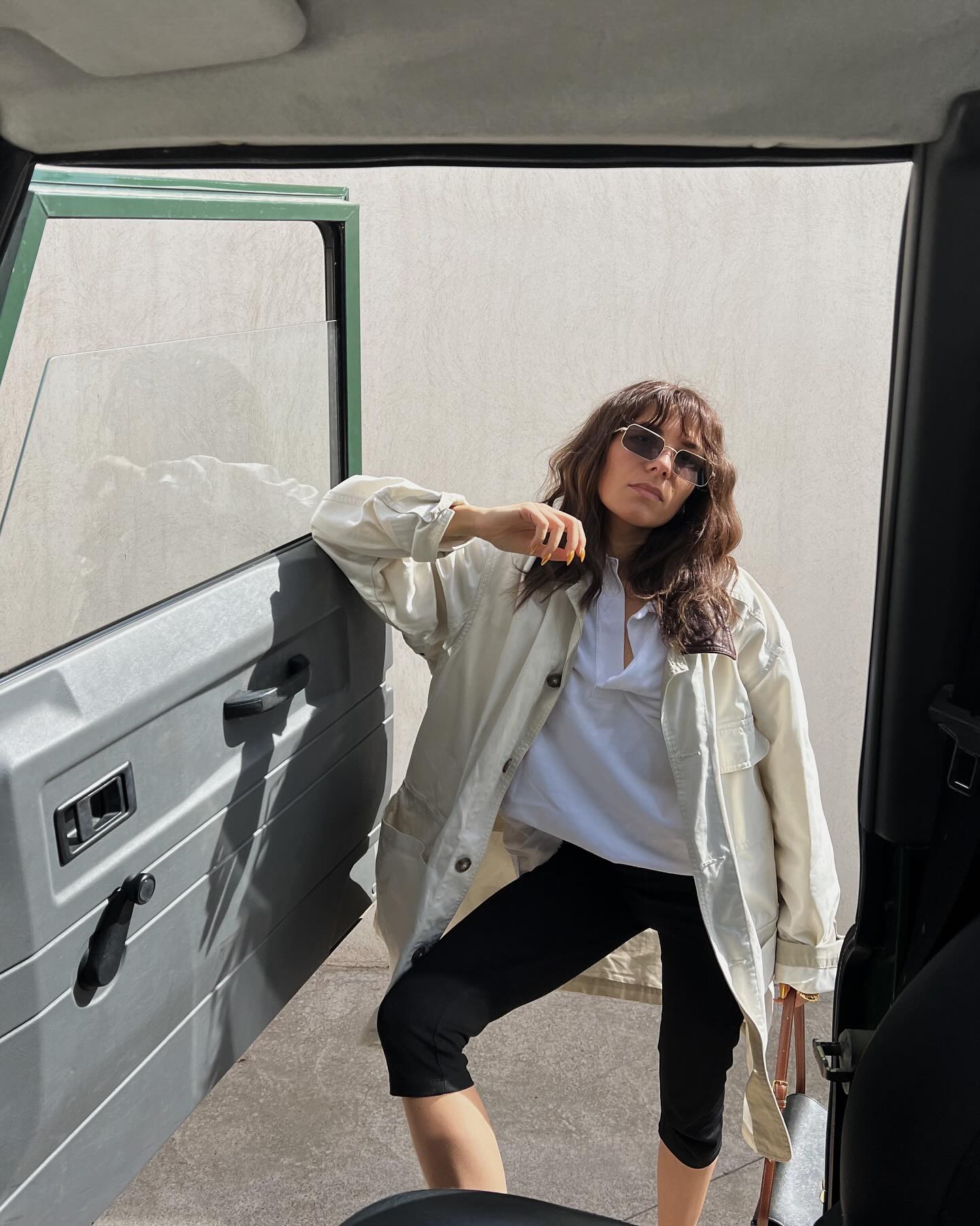 influencer Emilie T. poses in a car door frame with one leg up wearing wire-rim rectangular sunglasses, a barn-style chore coat, white button-neck top, and black capri pants