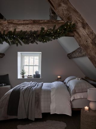 Christmas bedroom decor with garland on a beam, gray bedding, rug, basket style side table, lamp, mini Christmas tree