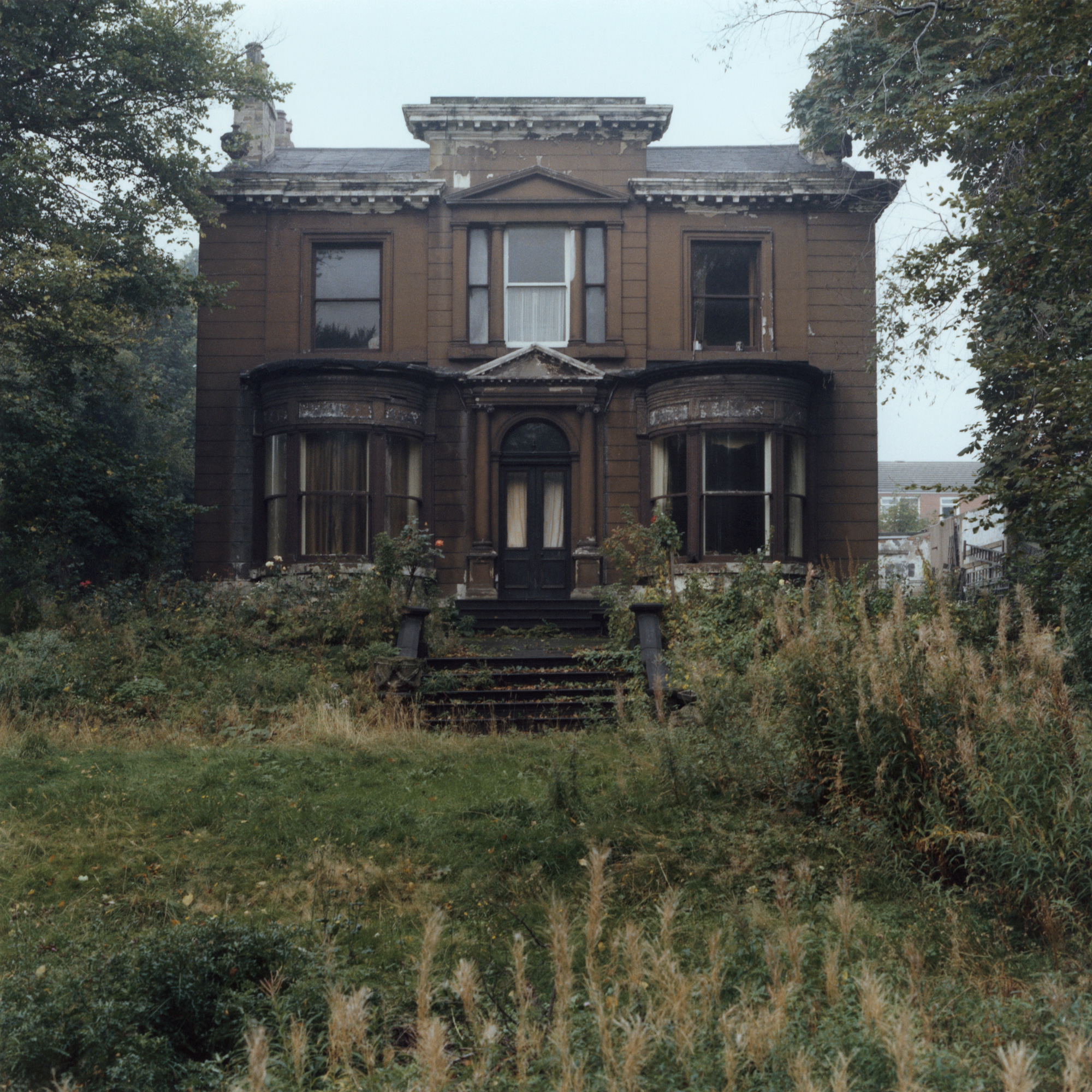 Colour photograph of a grand but decaying Victorian villa, its windows boarded or curtained, surrounded by overgrown grass and weeds.