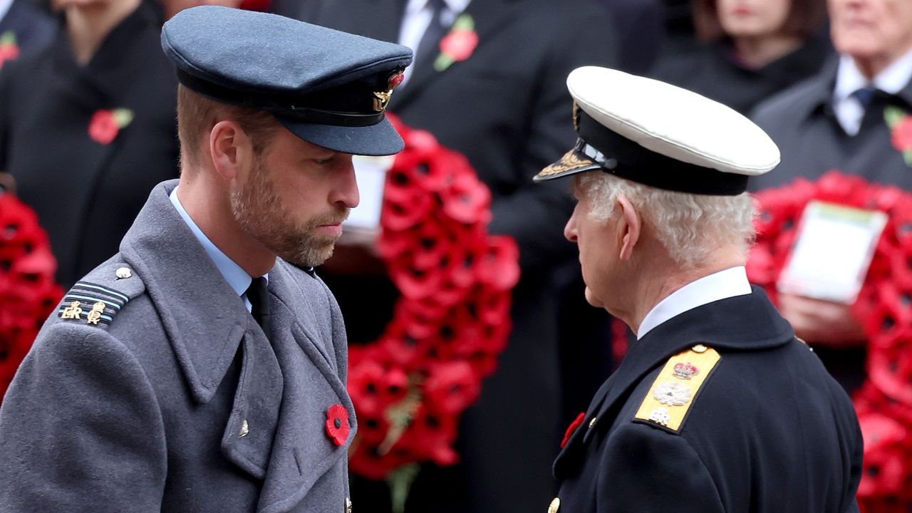 Prince William and King Charles facing each other wearing military uniforms in front of a wreath of poppies