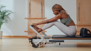 A woman sits on a Pilates reformer machine in a white room with a leafy plant.