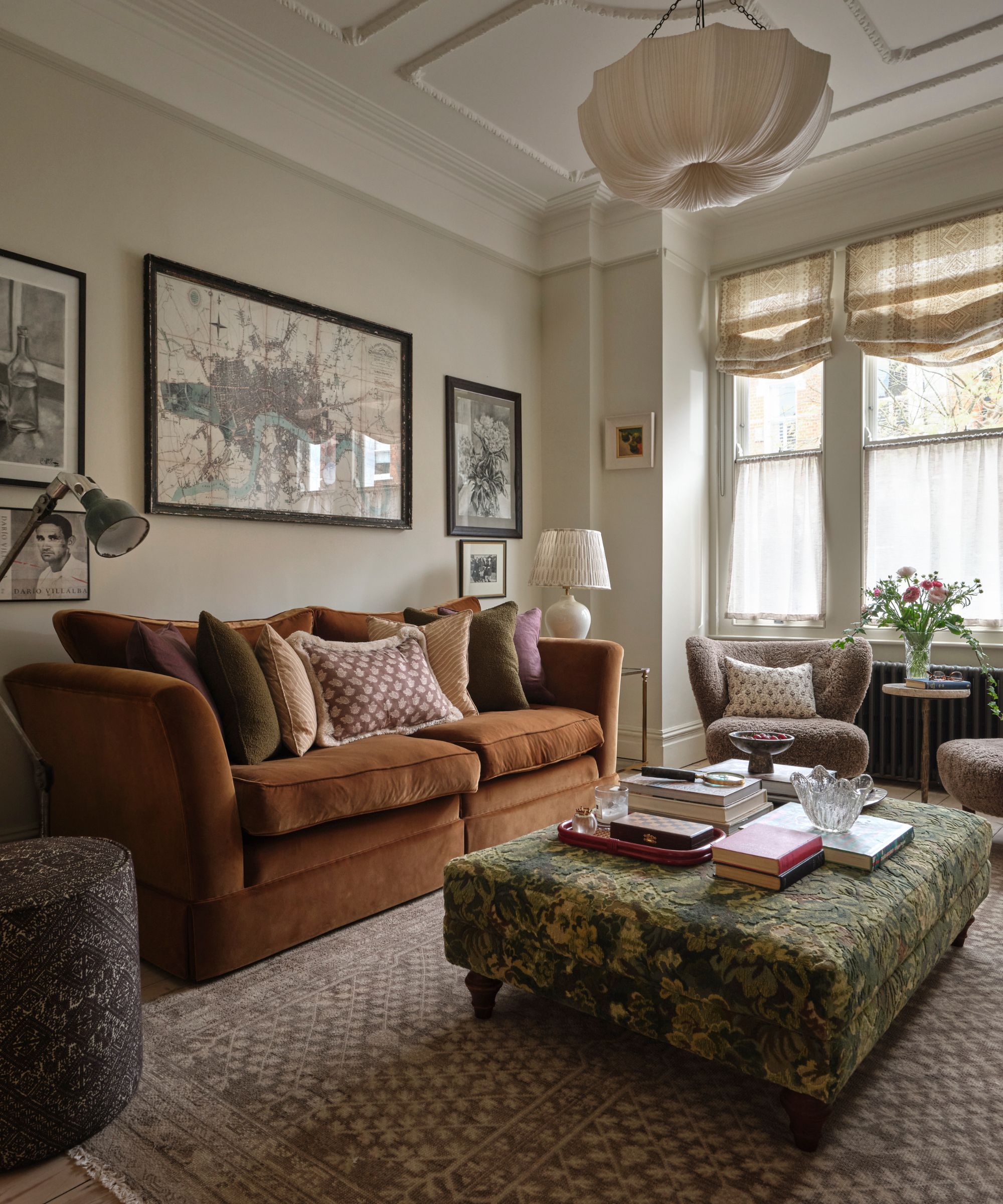 Living room with a rust velvet sofa, a jacquard ottoman with books and decor on top, an antique map print above the sofa, two boucle armchairs, and a pleated flush-mount light.