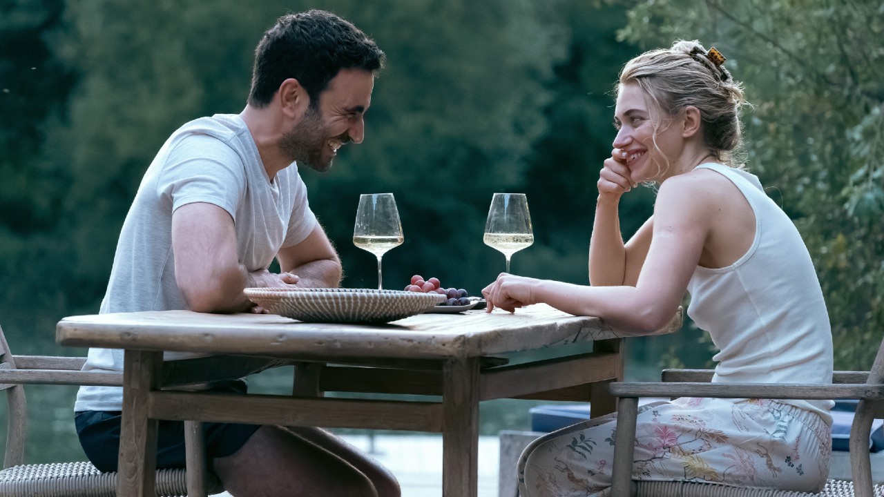 From left to right: Brett Goldstein smiling at Imogen Poots who is smiling back at him as they sit across from each other at a table with wine glasses next to them.