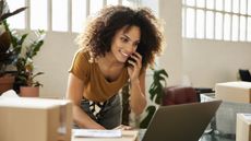 A woman smiles while on the phone and looking at her laptop while surrounded by boxes of inventory.