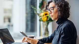 do blue light glasses actually work? - woman looking at her phone with black rimmed glasses on in a coffee shop with her laptop next to her