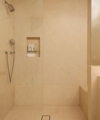 A beige walk-in shower with a glass door, a silver shower head, and a recessed shelf with toiletries in it