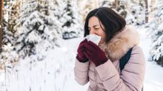 A woman is shown in a snowy forest. She is wearing a thick winter coat and gloves and is blowing her nose into a tissue. 