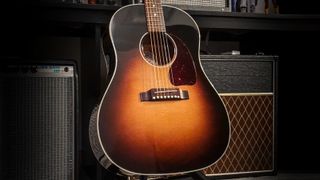 A Gibson J-45 acoustic guitar on a stand with various electric guitar amplifiers in the background