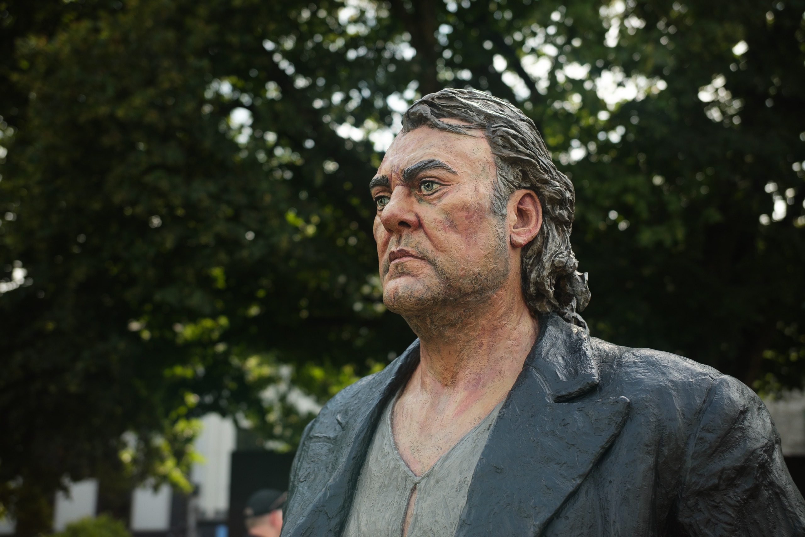 Close-up portrait of a painted outdoor statue of a man wearing a dark jacket, with trees in the background.