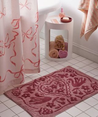 A bathroom with a pink rectangular patterned rug with a heart and plants on, a light pink bow shower curtain, a white side table with rolled towels underneath, and a hanging pink towel