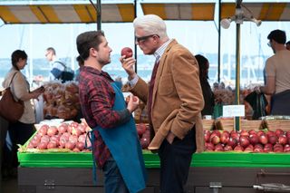 A man (Ted Danson as Charles, right) holds a peach as he stands uncomfortably close to a farmer's market vendor (left) in front of an outdoor display of peaches, in episode 101 of 'A Man on the Inside.'