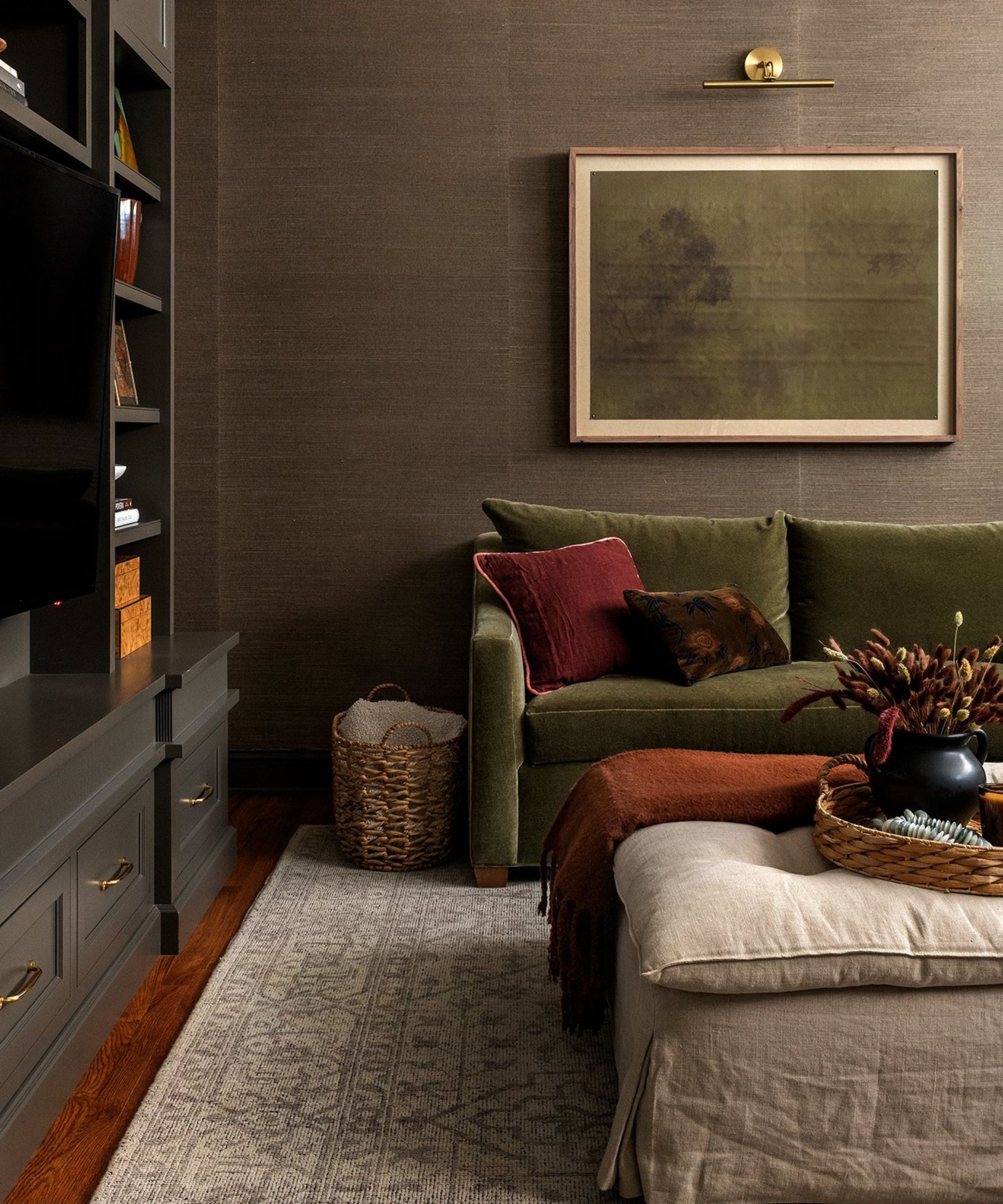 Dark living room with patterned rug, green sofa with cushions, framed minimalist painting, partial black bookshelf to the left, footstool, wooden floors.