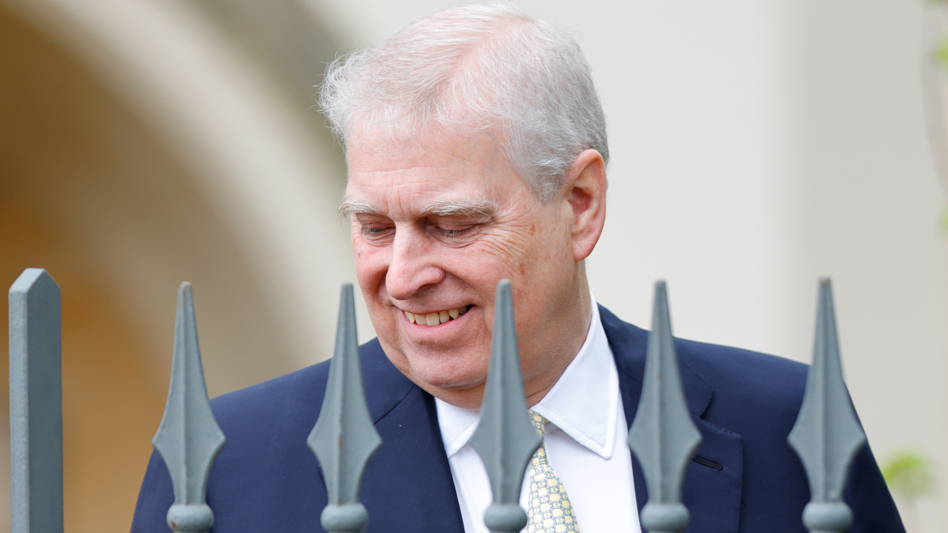 Ex-Prince Andrew wears a navy suit and stands behind a metal fence while smiling
