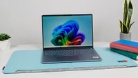 A sleek laptop on a light blue mat, displaying a colorful abstract wallpaper. It's flanked by a small plant on the left and stacked books on the right.