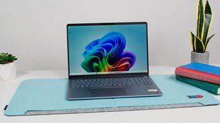 A sleek laptop on a light blue mat, displaying a colorful abstract wallpaper. It's flanked by a small plant on the left and stacked books on the right.