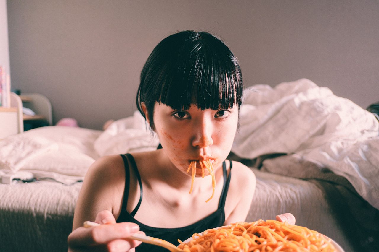 A girl holds a plate of spaghetti, with a bed and a simple backdrop suggesting a cozy indoor setting