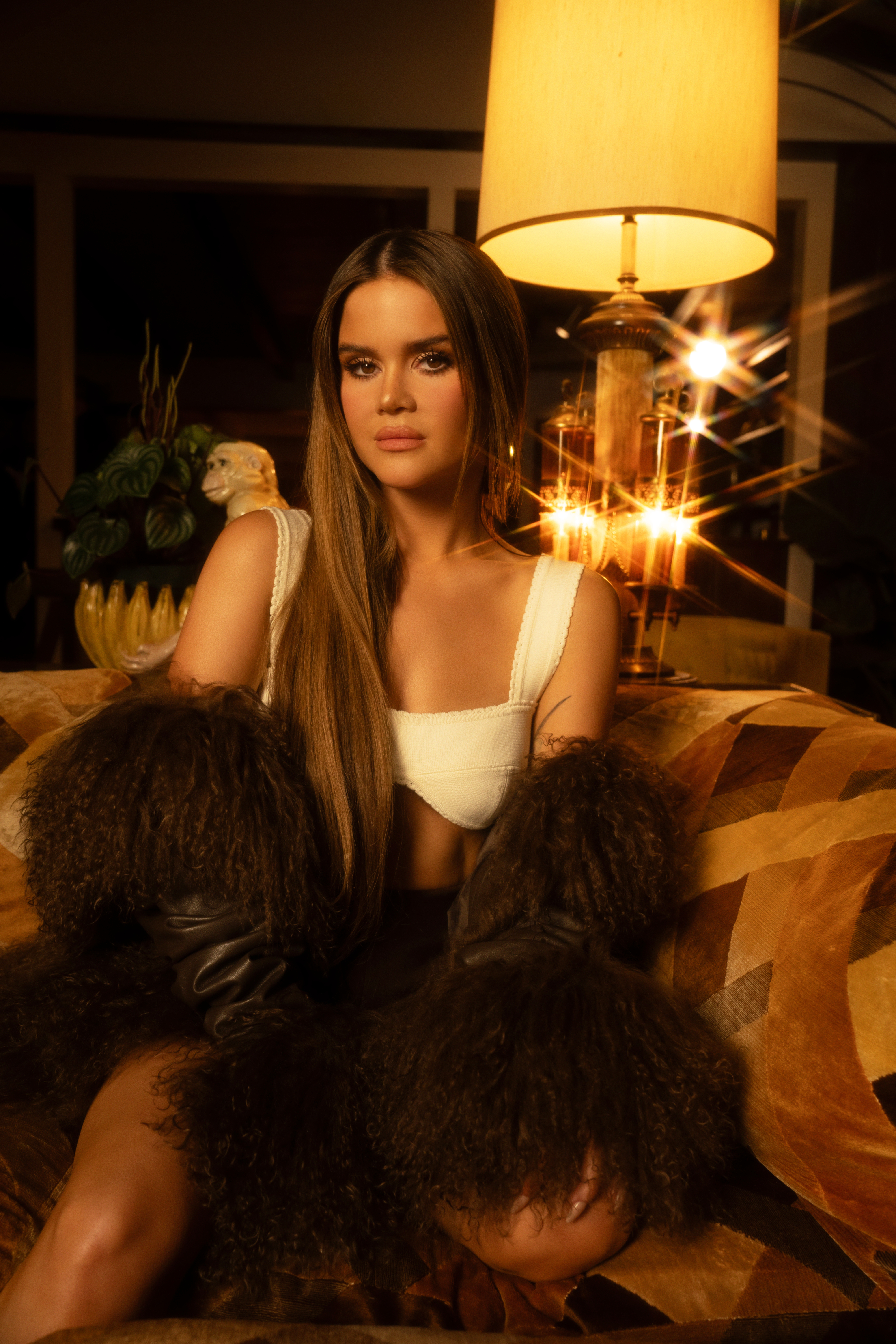 maren morris sits on a brown patterned couch wearing a white crop top and a brown fur coat