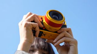TTArtisan AF 35mm f/1.8 II 'Orange Pop' on yellow and orange camera, held up to blue sky 