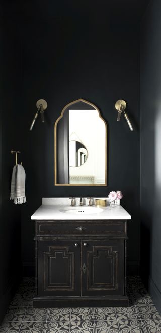 A gothic black bathroom with black patterned floor tiles, a gold mirror and gold wall lights