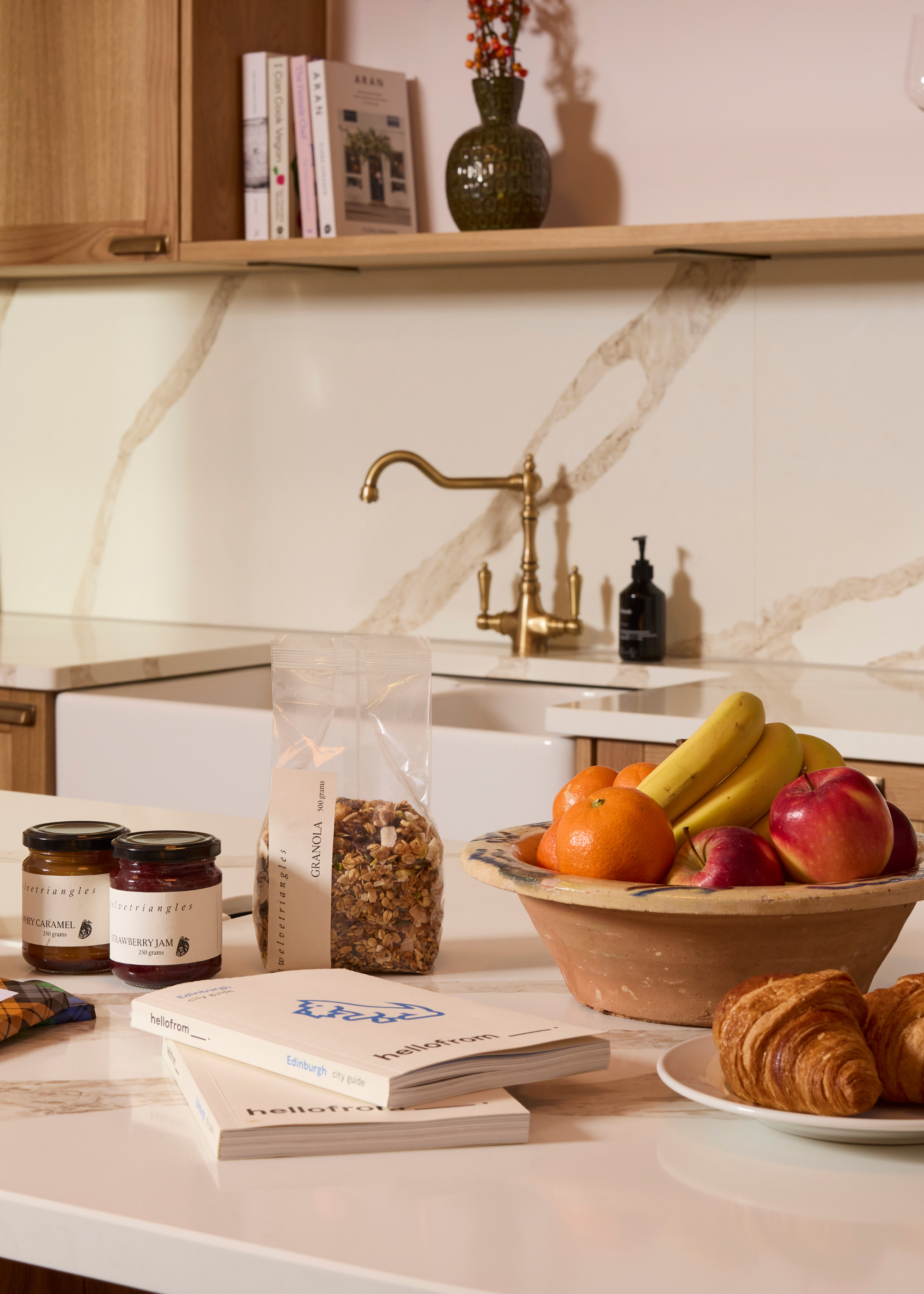 A stone kitchen island with a bowl of fruit, a plate of croissants, a couple of books, a bag of muesli, and a pair of jam jars