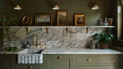 green and marble kitchen with sage green cabinetry, marble countertop, backsplash and shelf, green walls, marble sink, brass hardware, artwork on shelf, wall lights