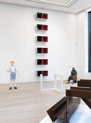 We see a sculpture of a boy in the back, with a sculpture of a delivery man. White shelves with red & black inside are stacked vertically on the wall.
