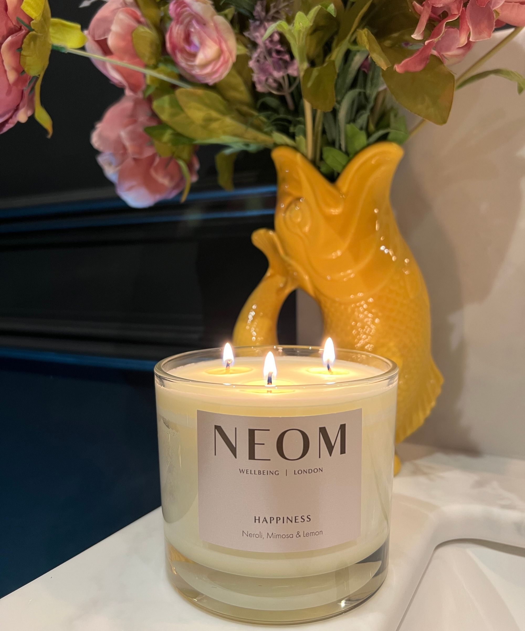 A lit NEOM Happiness Candle on a white marble sink with a yellow fish vase filled with flowers and a black panelled wall behind.
