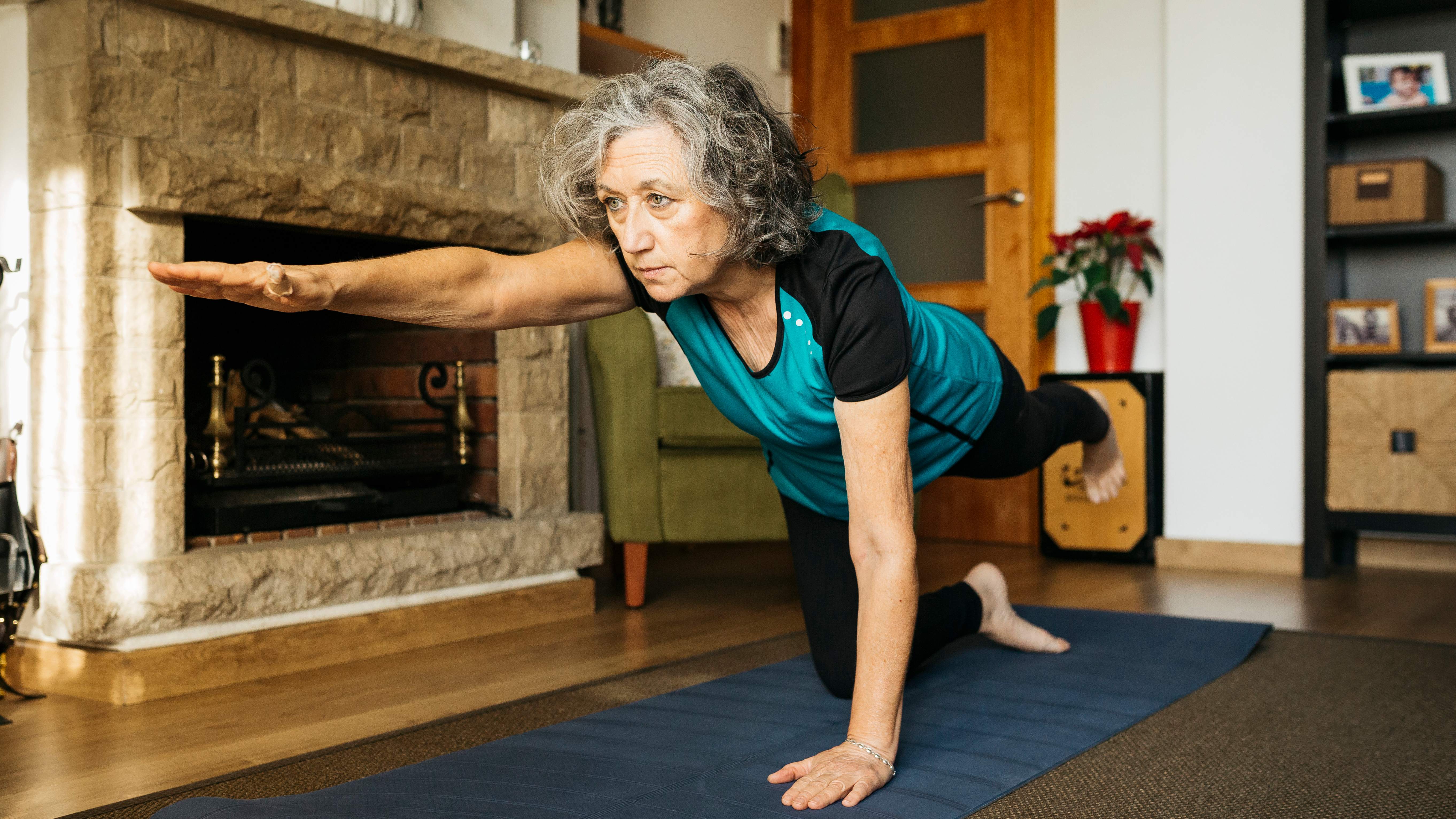 A woman performs bird dog exercise at home. She is balancing on her right knee and left hand, with her torso parallel to the floor, and her right arm extended forward, left leg extended behind her.