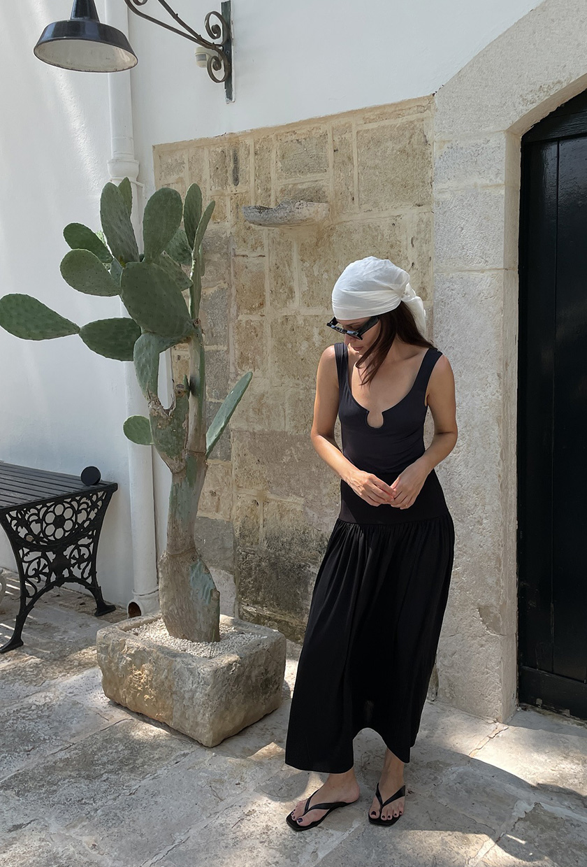 The coastal boho trend shown in a street style photo of a woman standing outside a resort near a cacti wearing a white silk headscarf with black sunglasses, a black flowy boho maxi dress, and black flip flops